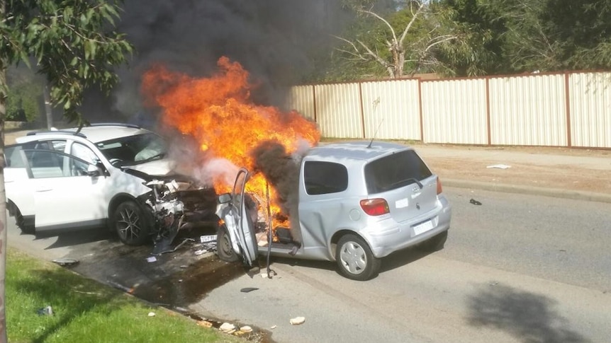 Two cars in the middle of a road, smashed head on into each other with flames and smoke.