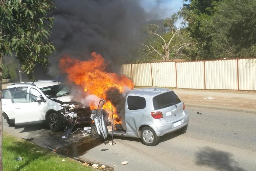 Two cars in the middle of a road, smashed head on into each other with flames and smoke.