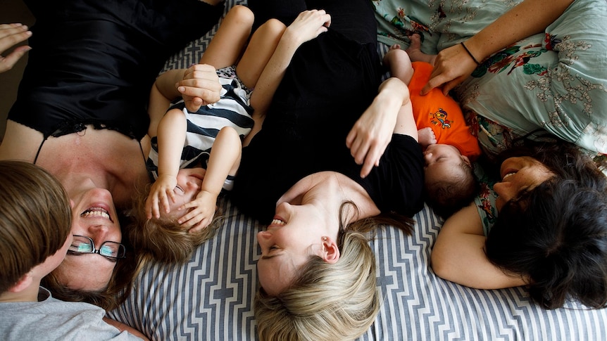 Three women lie on a bed with a baby and a toddler for a story on share housing
