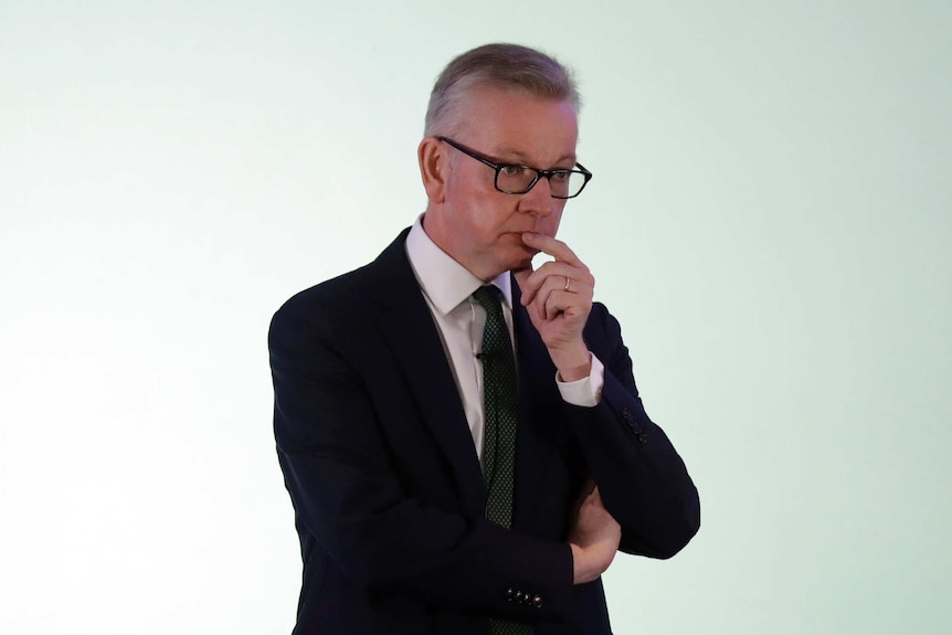 Michael Gove wears a suit and appears to be pondering as he stands in front of a wall