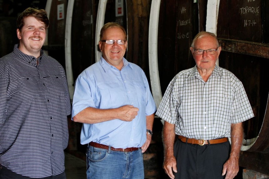 Three men stand in front of wine barrels