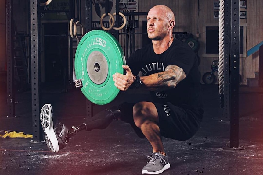 Paul is squatting on one leg and holding a weight plate in a gym