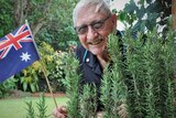 Man with glasses holding Australian flag and smiling in front of rosemary bush