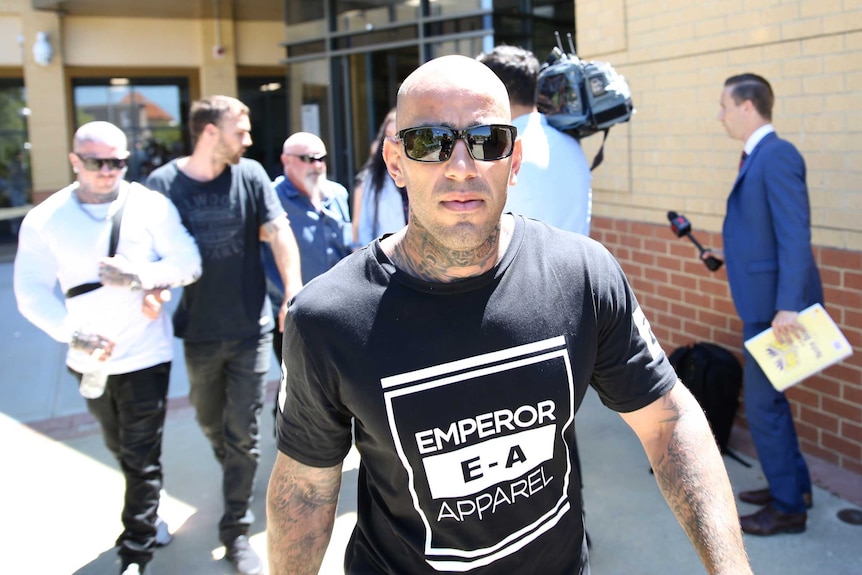A heavily tattooed man in a dark t-shirt and sunglasses in front of a crowd of people outside a court house.