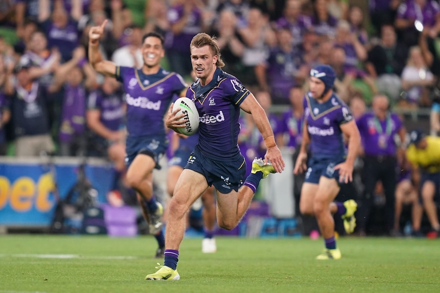 Rugby league player running towards the try line with his teammate celebrating in the background.