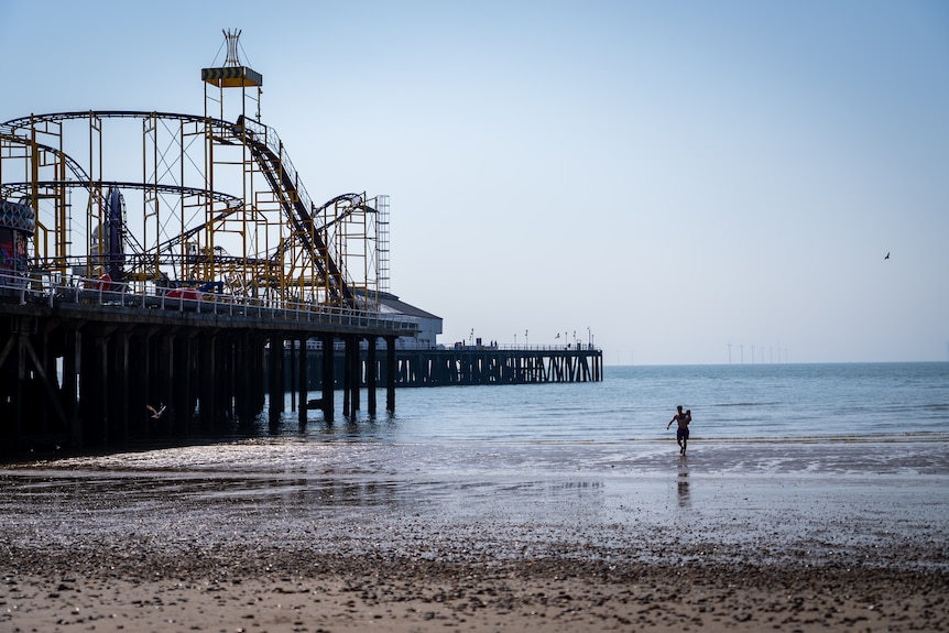 An empty rollercoaster and beach with receding tide sits against a stark blue sky with a man carrying the child toward the water