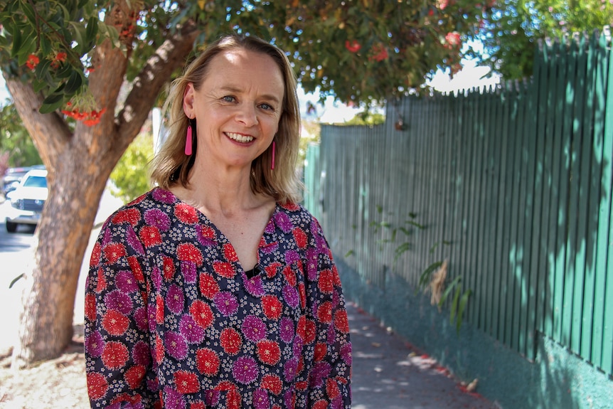 Nina Rogers stands on a footpath in front of a red flowering gum and smiles at the camera