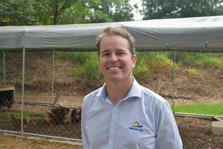 A man wearing a pale blue collared shirt smiles, he stands in front of a wire enclosure.