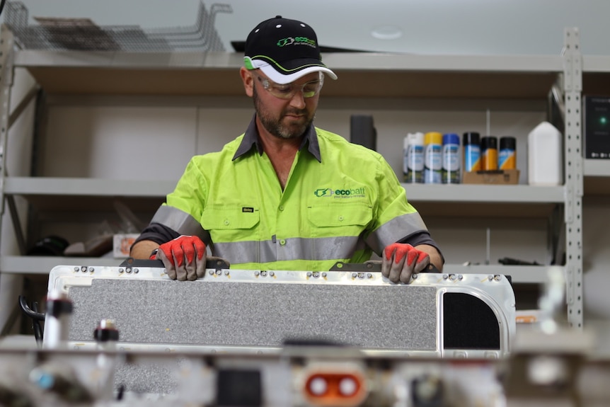 Hamish wears a black cap, goggles, yellow shirt and red gloves. He is holding a metal sheet. Industrial shelves are behind him.