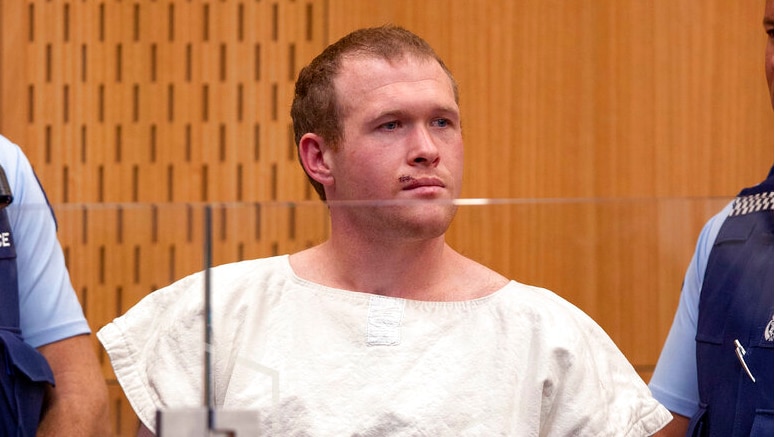 A man in a cream coloured prison uniform between two police officers