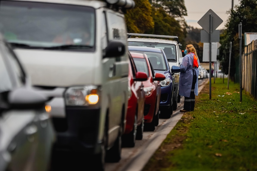 A woman in a medical gown and face mask and shield stands by a line of cars. 