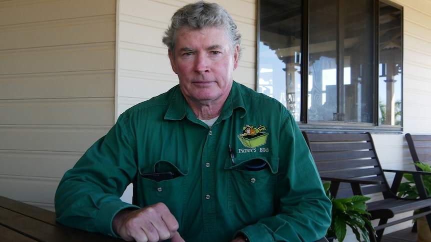 A man with grey short hair sits on a veranda in a wooden chair wearing a green shirt and looks at the camera.