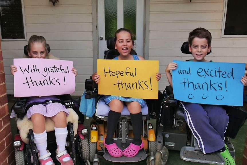A group of triplets with muscular dystrophy hold 'thank you' signs while sitting in wheelchairs.