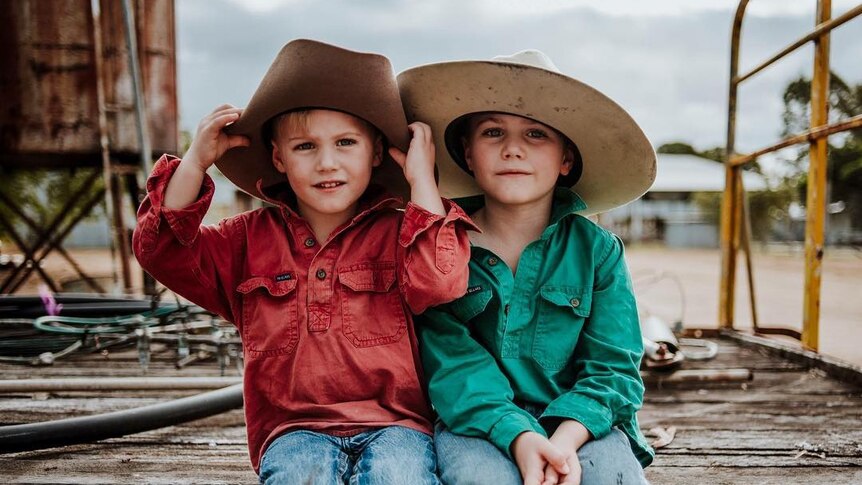 Two young boys wearing big hats sit on the back of an old hay cart.