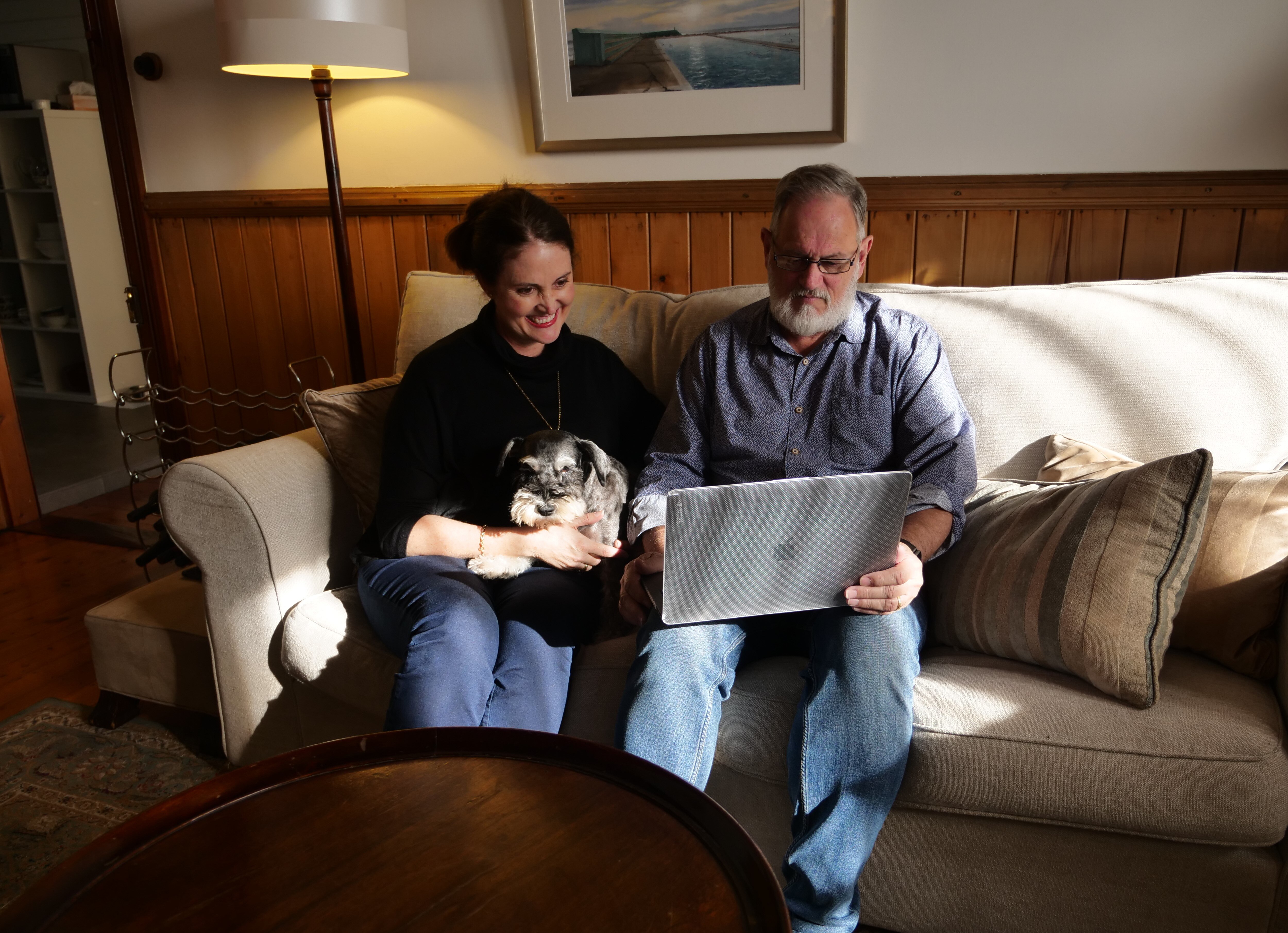 A man and woman, the latter with a cat on her lap, sit on a couch looking at a laptop.