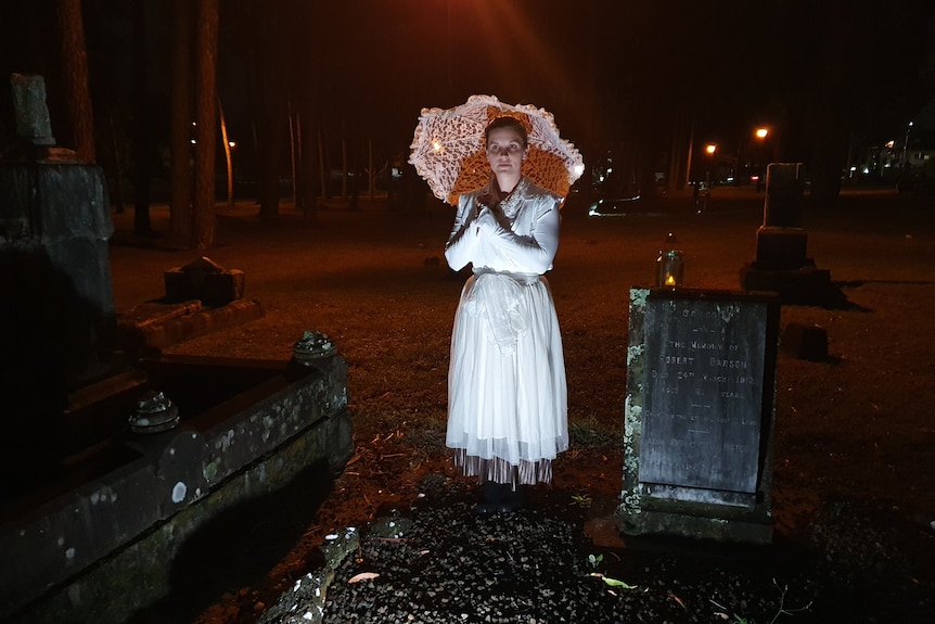 Dressed in a white period costume and standing beside a grave, woman holds umbrella.Spotlight on her, darkness behind.