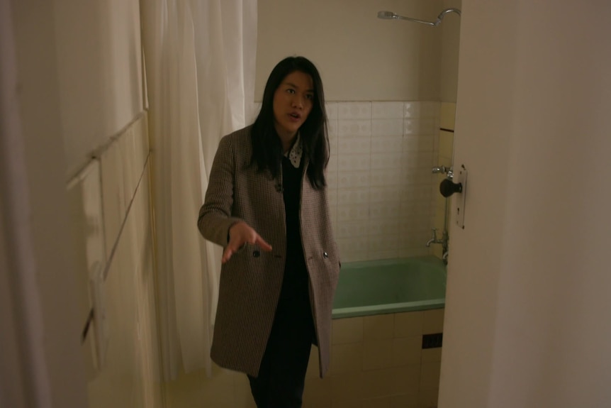 Angelique stands in a dated bathroom with a green bathtub