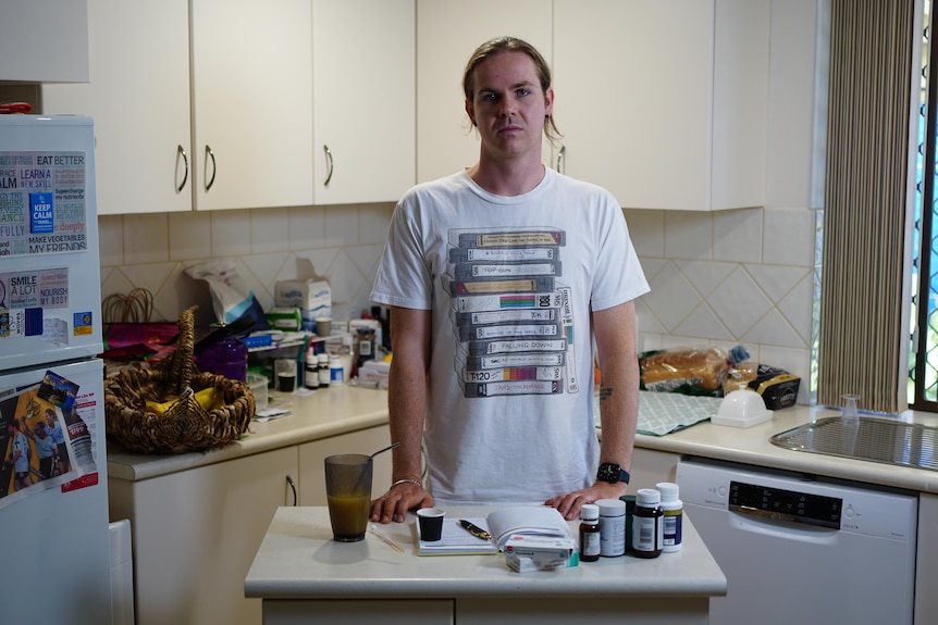 A young white man with long brown hair standing in a kitchen next to an array of medicine bottles
