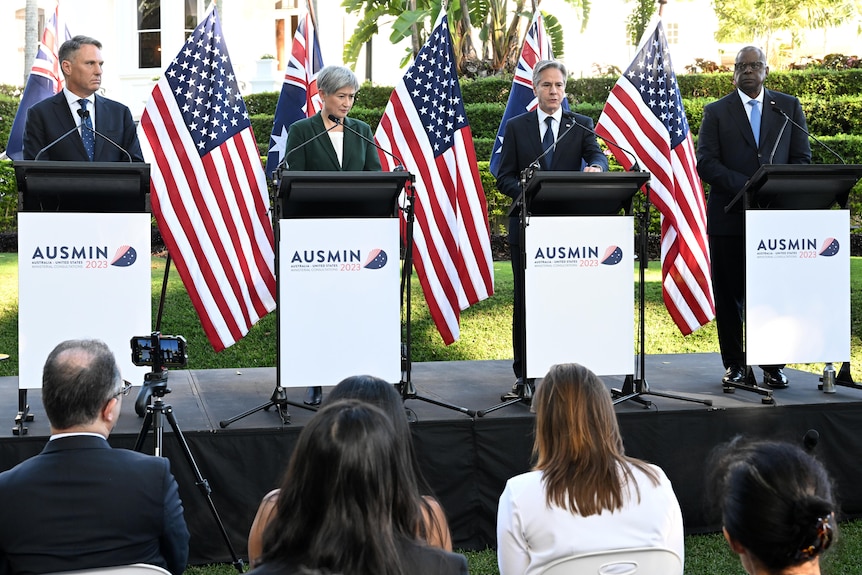 Four dignitaries in suits, three men and one woman, stand on podium in front of USA and Australian flags