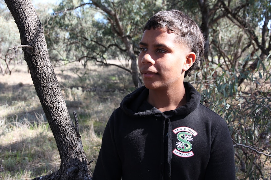A teenager in dark, warm clothing stands in bushland.