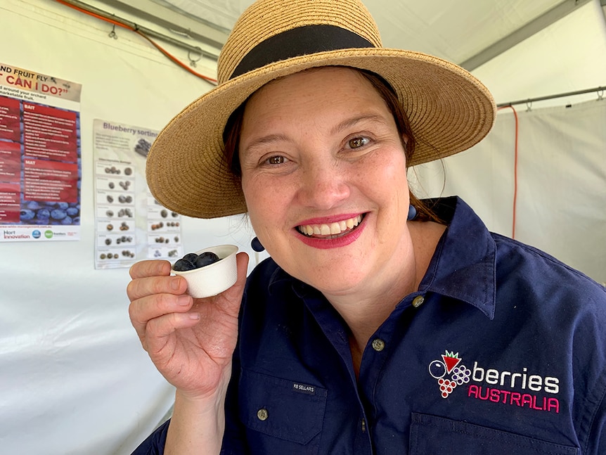 A woman wearing a straw, broad-brim hat smiles while holding a small container of blueberries