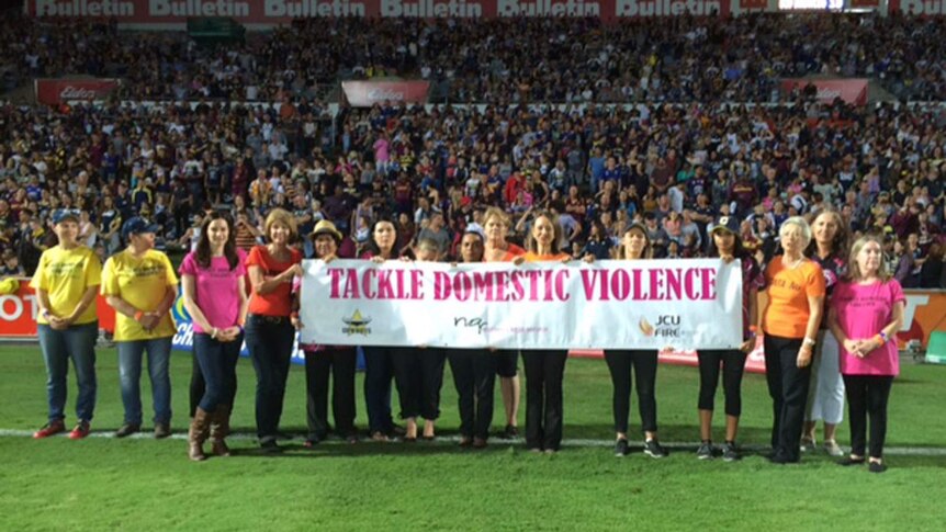 A group of people wearing bright t-shirts hold a banner reading 'tackle domestic violence' while in a sports stadium