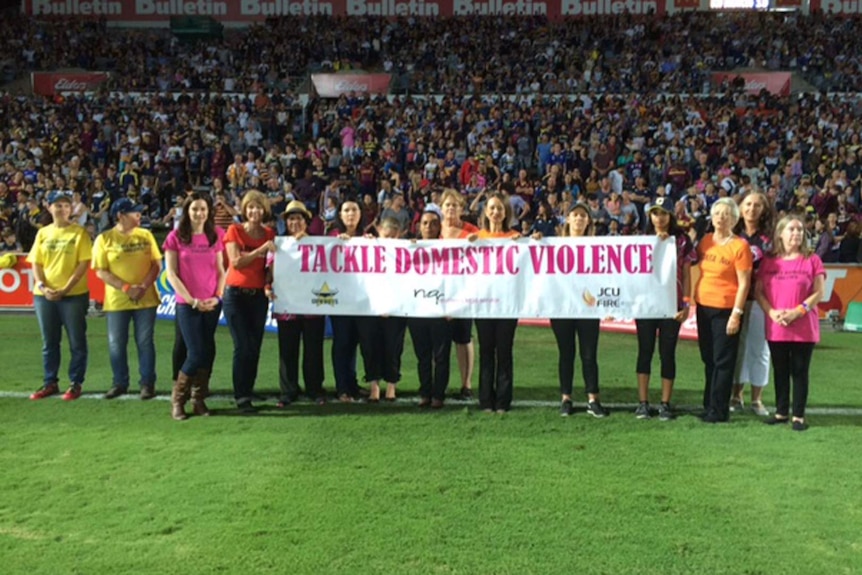 A group of people wearing bright pink t shirts hold a banner reading tackle domestic violence' while in a sports stadium