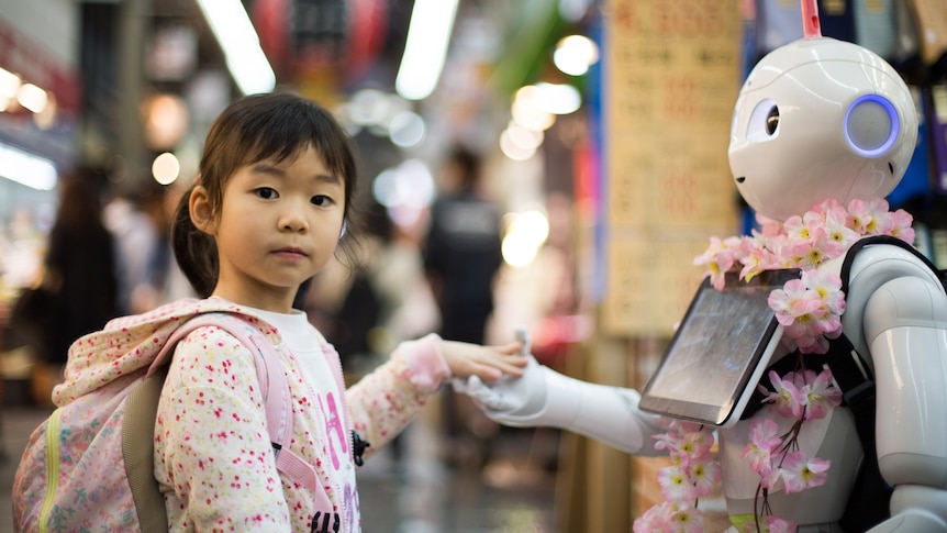 A young girl wearing a backpack looks at the camera while shaking the hand of a robot slightly taller than her