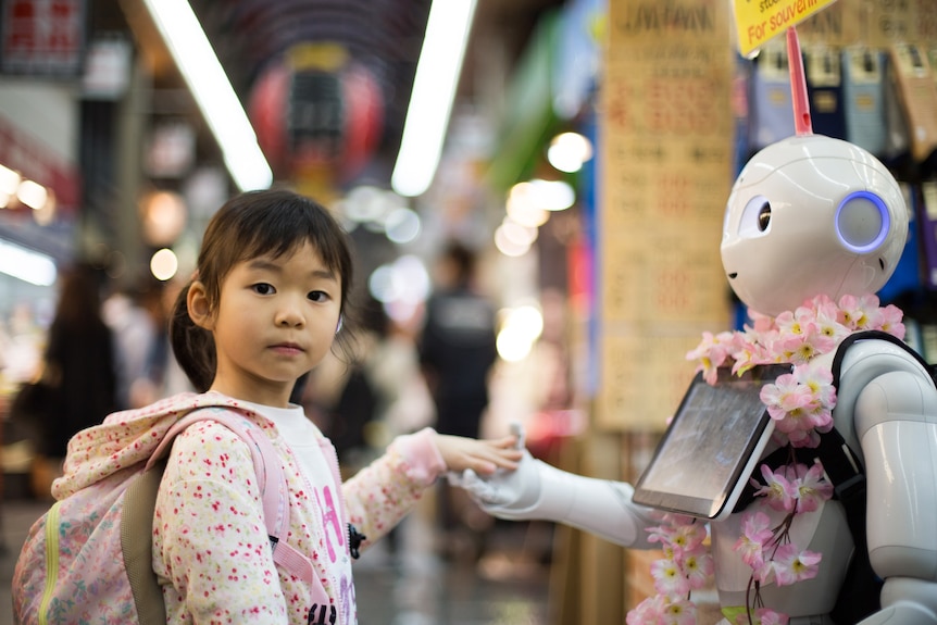 A young girl wearing a backpack looks at the camera while shaking the hand of a robot slightly taller than her