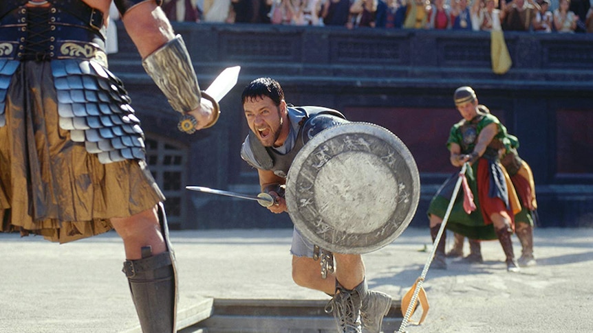 Russell Crowe acts as a gladiator in the arena charging towards and enemy holding a sword and shield.