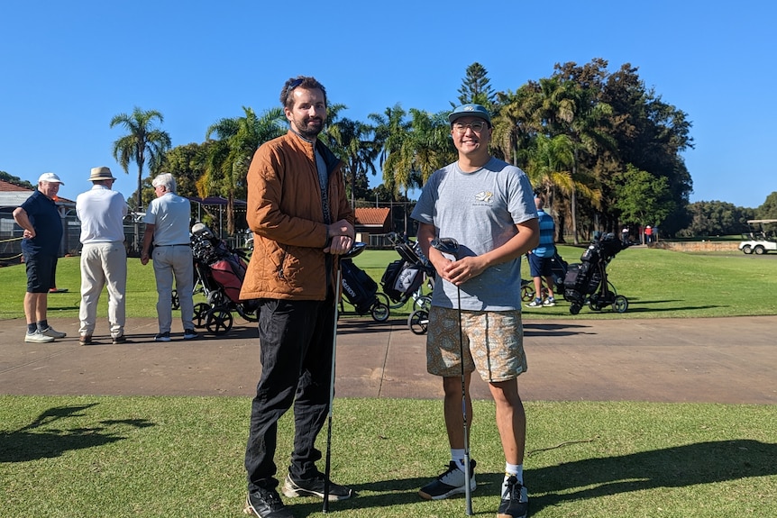 Dan Kohlman and Aaron Lee pose with the golf clubs and other golfers in the background 