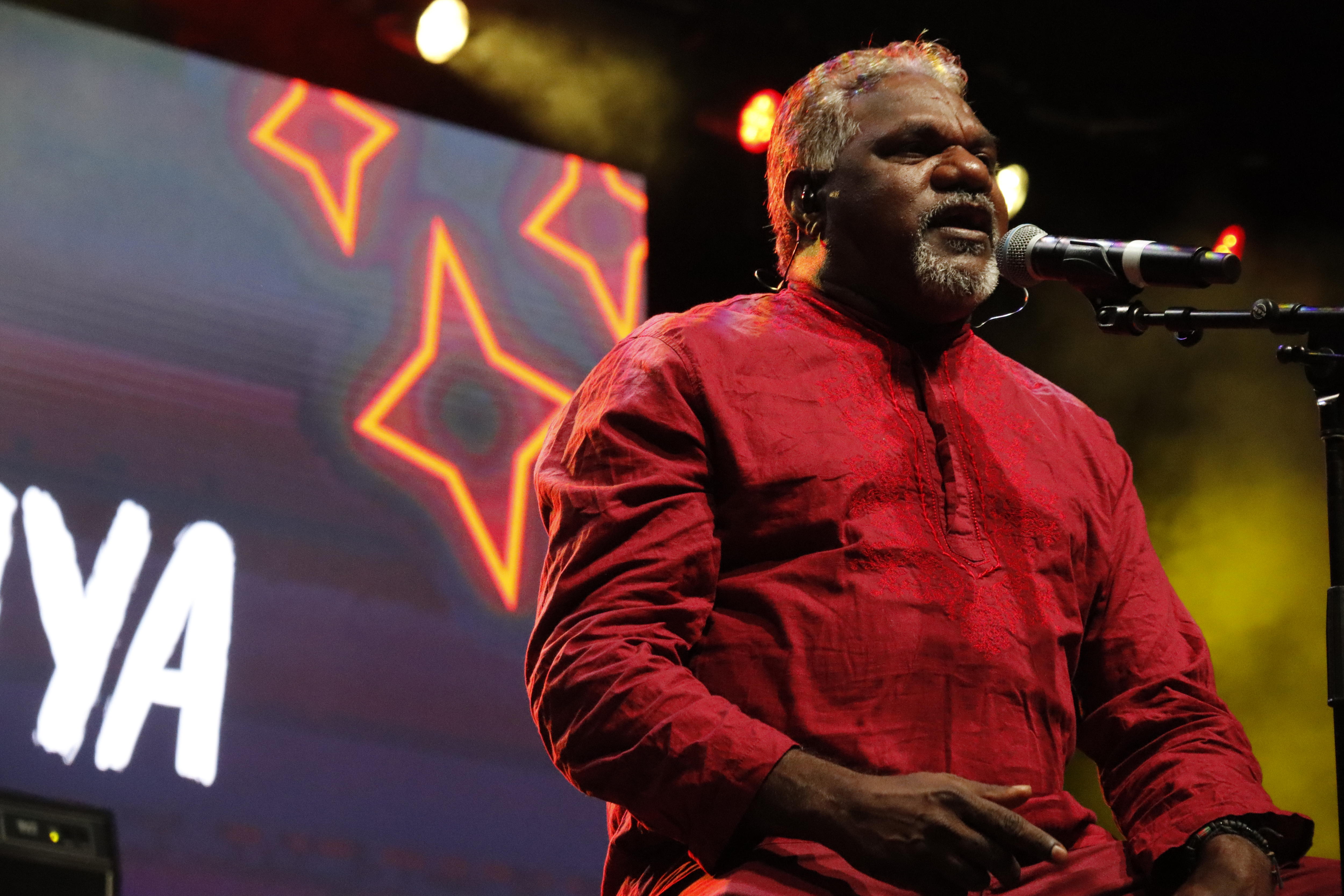An older Indigenous man sitting on a stool on a stage, singing into a microphone