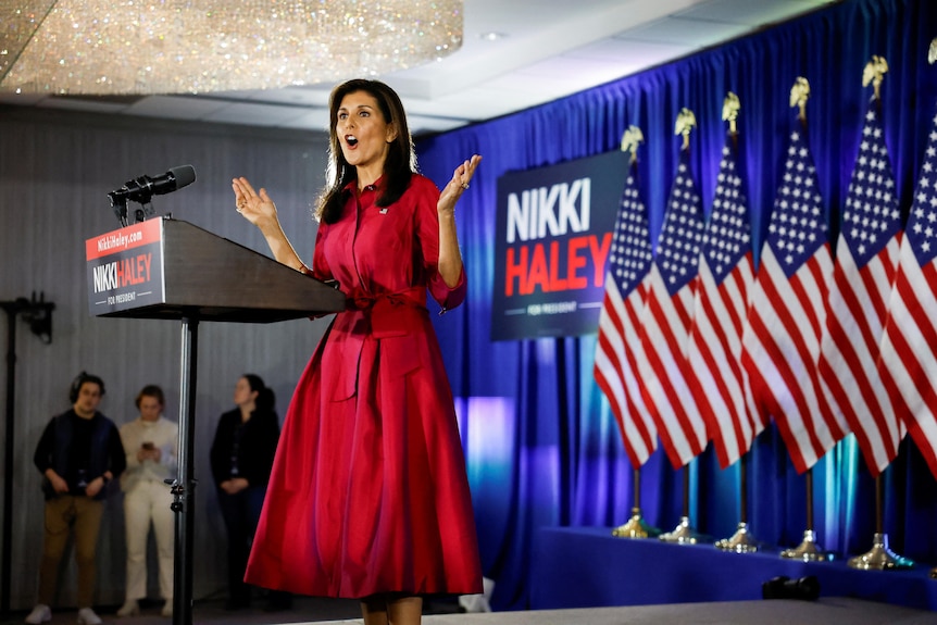 Nikki Haley, wearing a red dress, stands at a podium and holds her arms open. US flags are behind her.
