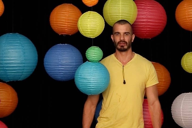 A bearded man in a yellow t-shirt stands among colourful hanging lanterns.