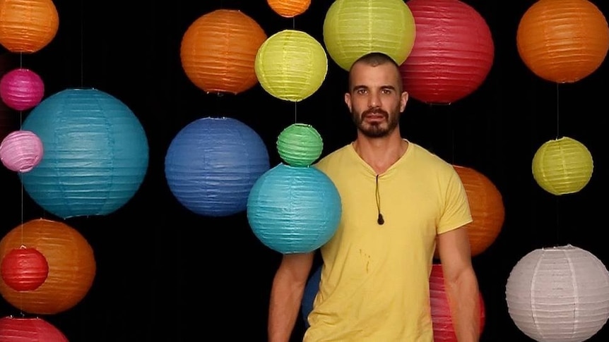 A bearded man in a yellow t-shirt stands among colourful hanging lanterns.