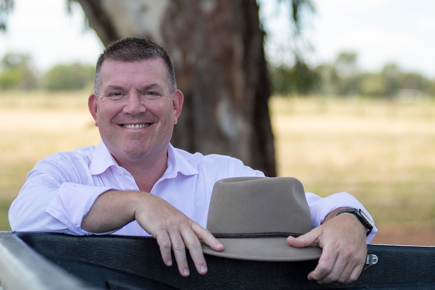 A man with short hair stands in front of a tree and smiles at the camera. He is holding a wide-brimmed hat