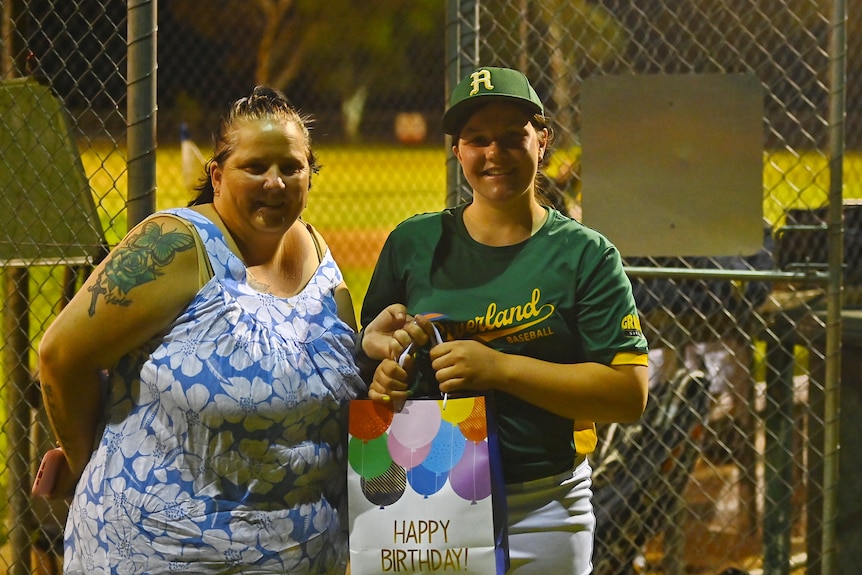 A smiling woman and a girl in a baseball uniform. She holds a bag with "happy birthday" written on it.