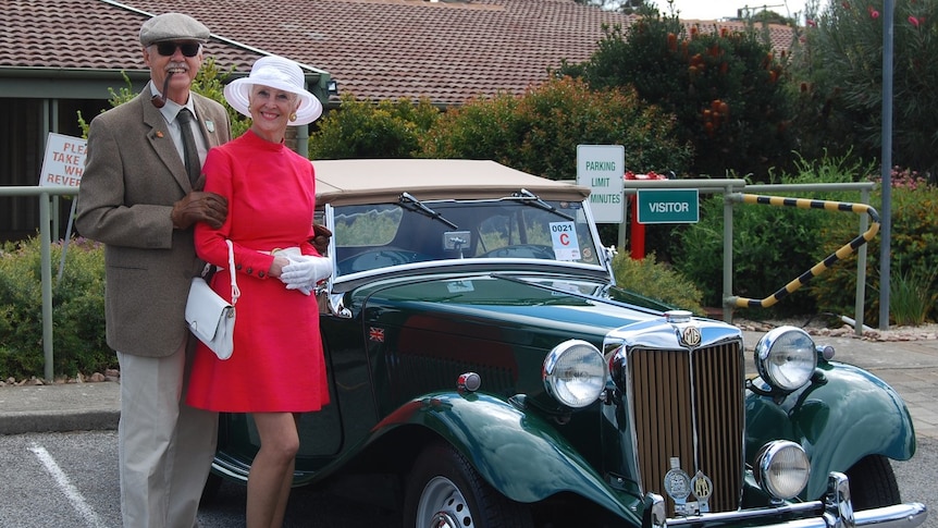A woman in a dress and a hat stands next to a man in a suit with a pipe beside an old car with big round lights.