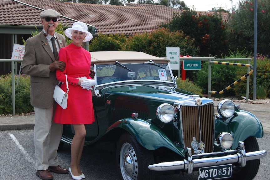 A woman in a dress and a hat stands next to a man in a suit with a pipe beside an old car with big round lights.