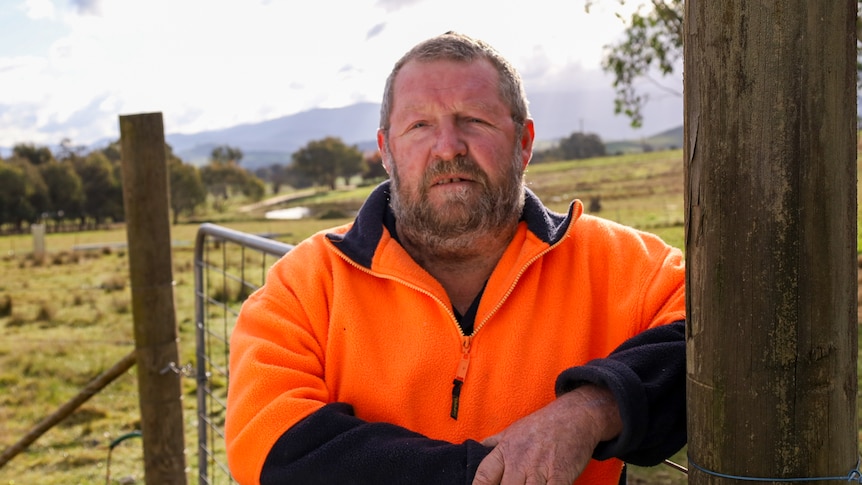 Man in orange fluro jumper leans against fence, with open farmland in the background. 