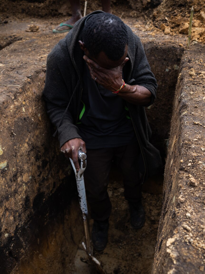 A man standing in a dug out grave in the dirt, holding a shovel, with his head down and hand on eyes