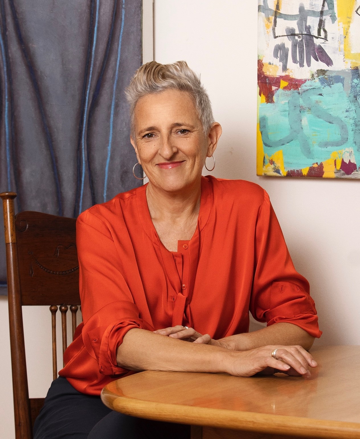 A woman with short grey hair and a half smile ,wearing an orange top, sits at a table