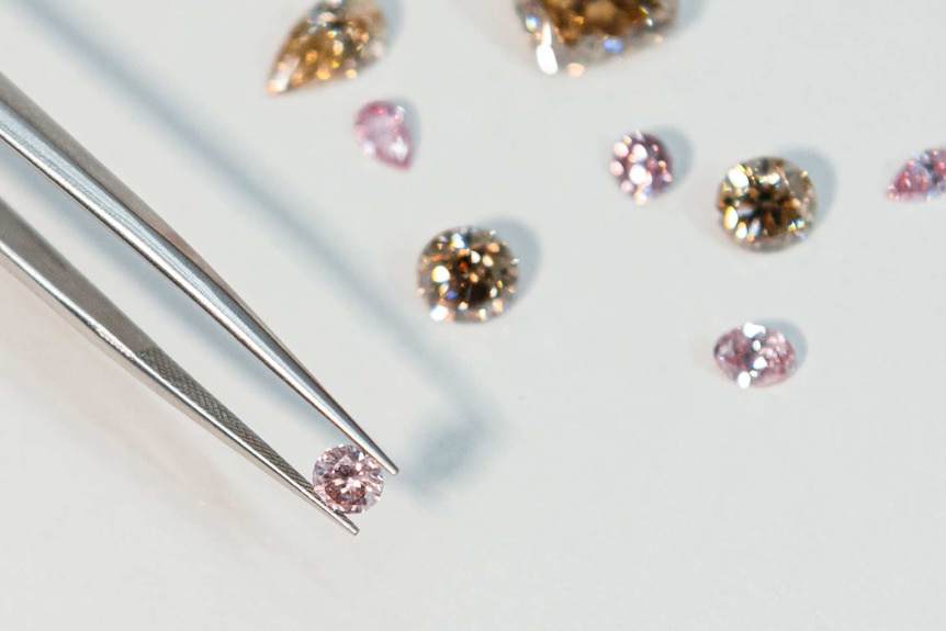 A selection of coloured diamonds on a white surface with one pink diamond held by metal tweezers.