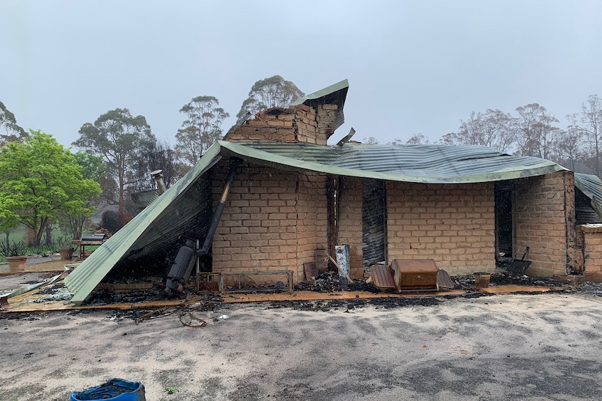 A collapsed brick house with a corrugated iron roof sits on sandy ground on a grey day.