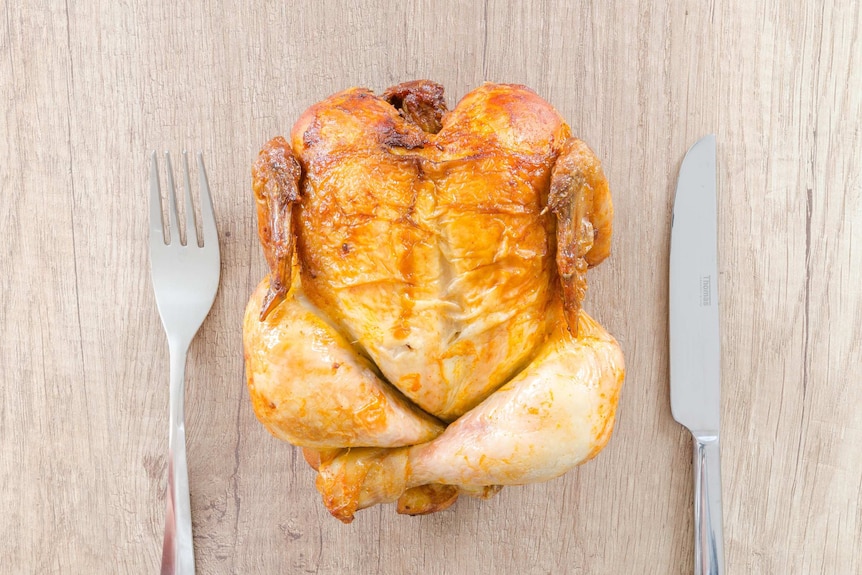 A roast chicken sits on a table next to knife and fork.