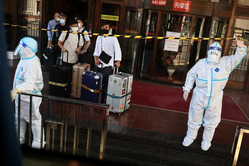 People walk out of a hotel with luggage past police tape and staff in PPE