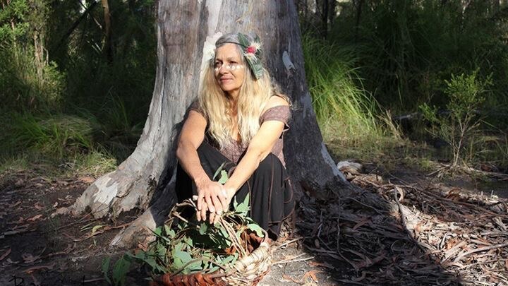 A woman with long blonde hair sits at the base of a tree, with a basket of leaves at her feet.