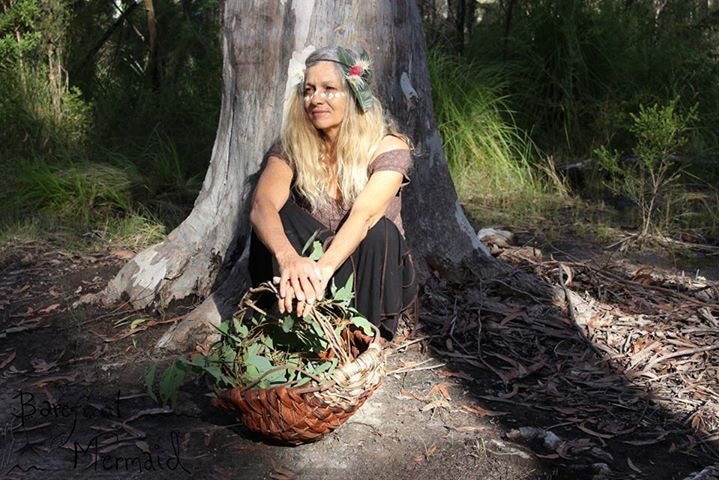 A woman with long blonde hair sits at the base of a tree, with a basket of leaves at her feet.