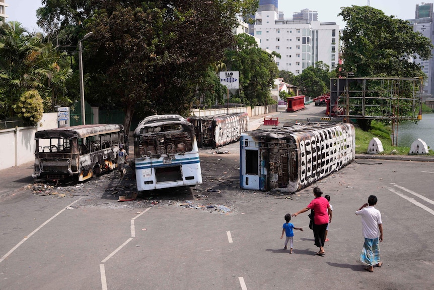 Four people look at burnt out buses lying on a road.