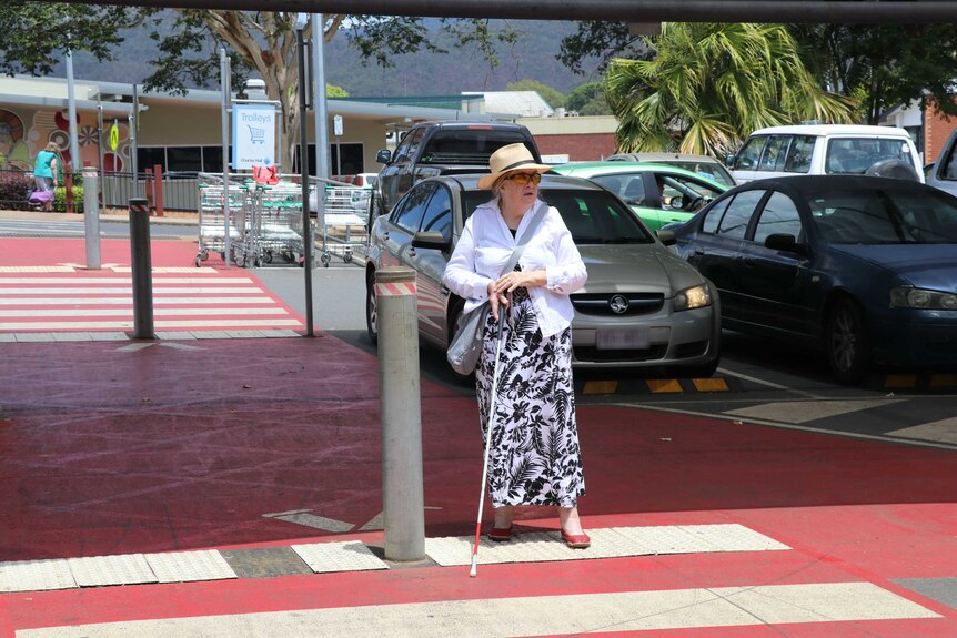 A woman with a white mobility cane walks on a pedestrian crossing in a carpark.
